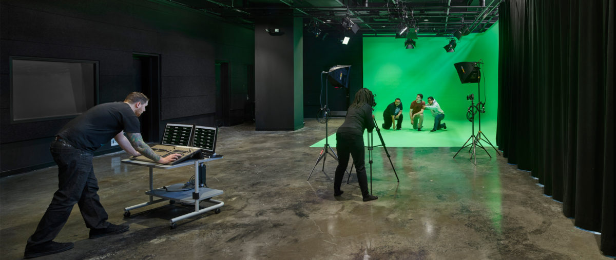 Two people film a group of three posing in front of a green screen.