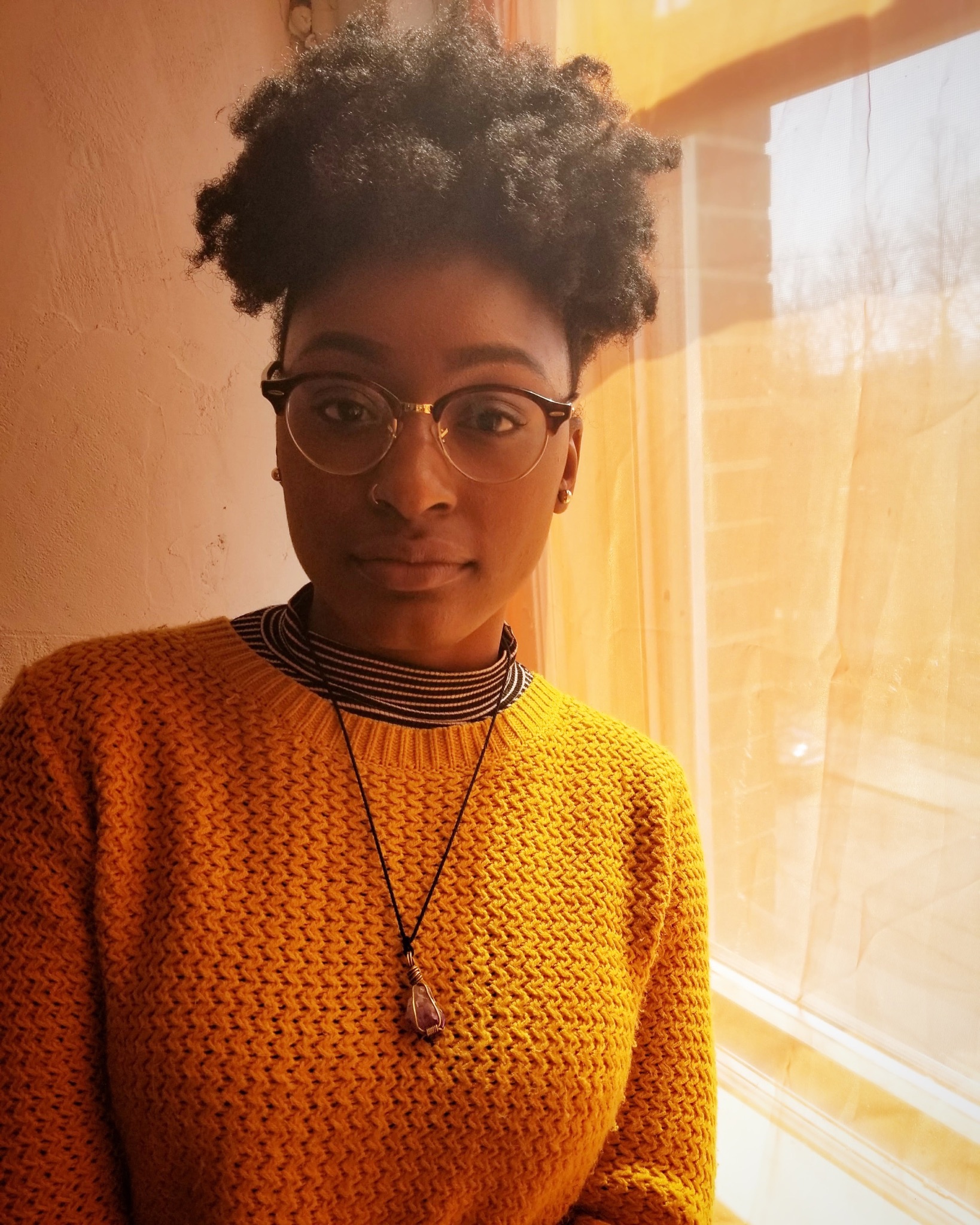 color photograph, headshot of Anika Carter in a yellow sweater leaning on a window and staring straight into the camera