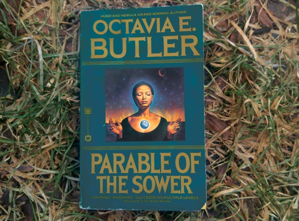 A book lies on the grass. The book covers shows a black woman with her eyes closed, a small globe of light floating in from of her chest. The title of the book is "Parable of the Sower" and its author is Octavia E. Butler.