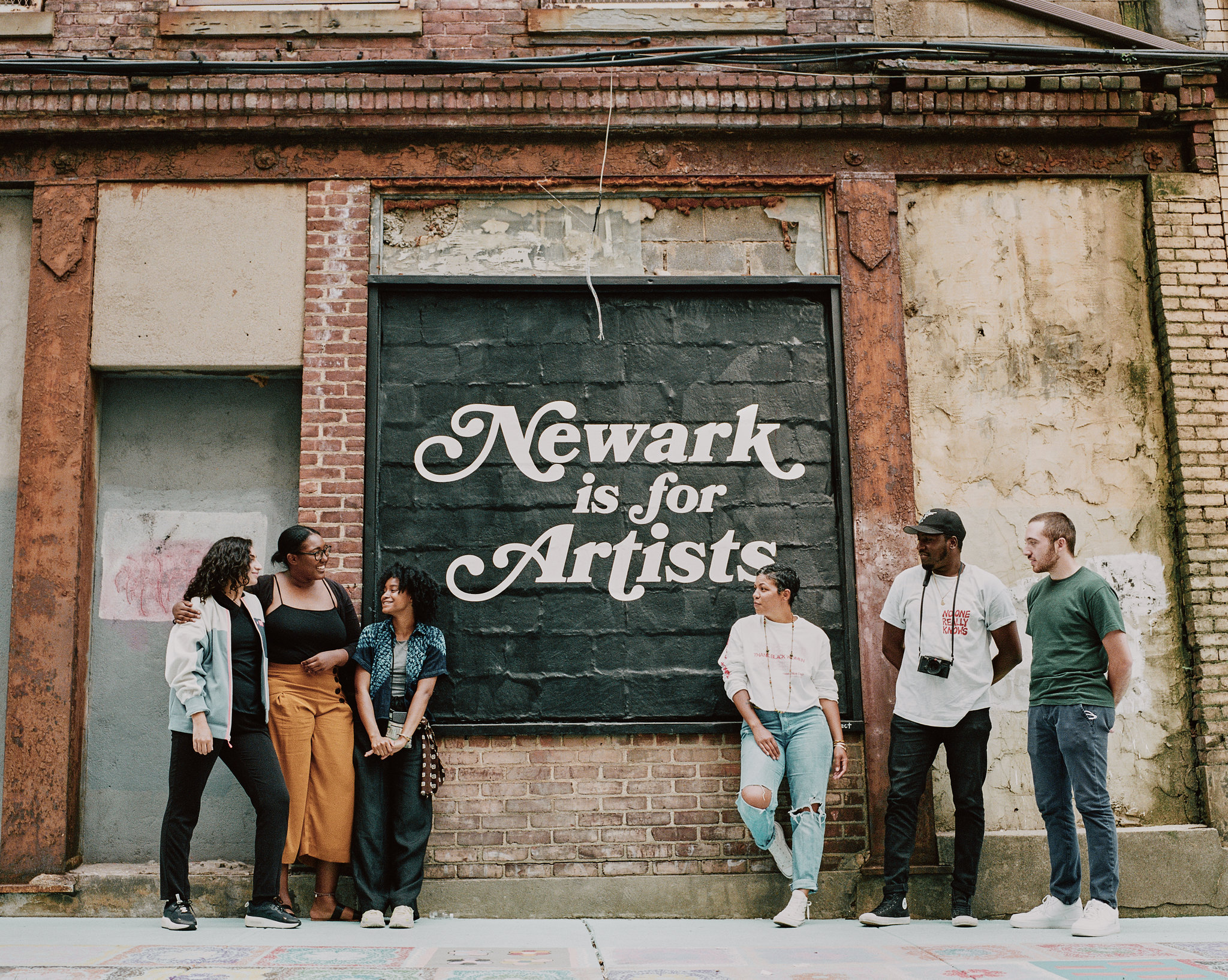 A group of people stand outside, in front of a mural that reads, "Newark is for Artists"