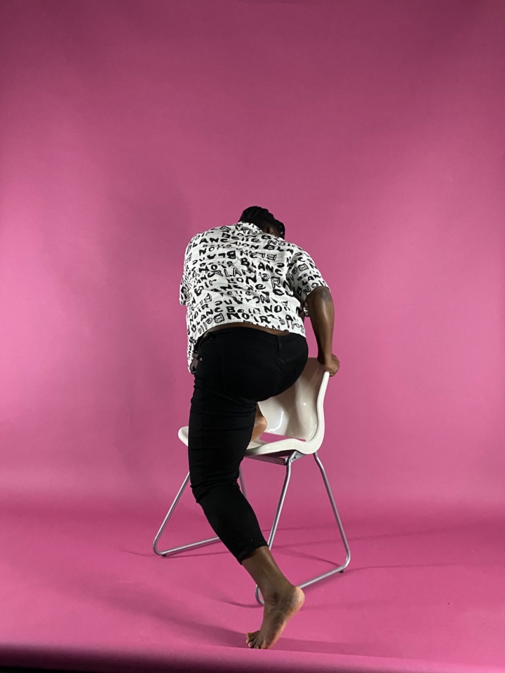 A Black woman, turned away from the viewer, climbs a white chair in front of a pink backdrop.